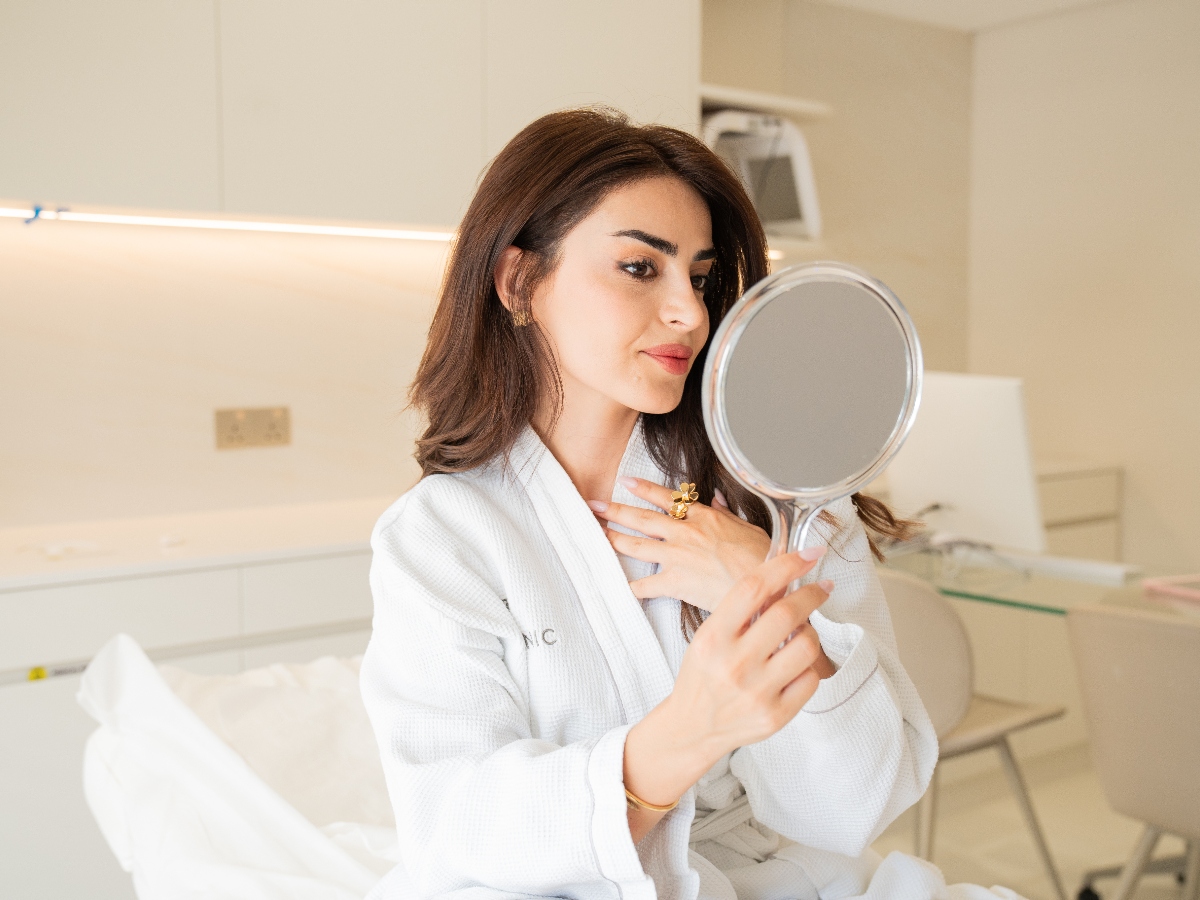 A woman in a white bathrobe examines her reflection in a handheld mirror, seated in a modern, minimalistic room.