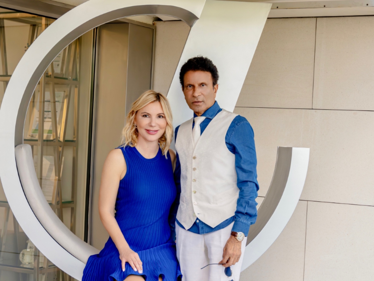 A man and woman pose by a circular sculpture. She wears a blue dress, and he wears a blue shirt and white vest. The atmosphere is stylish and composed