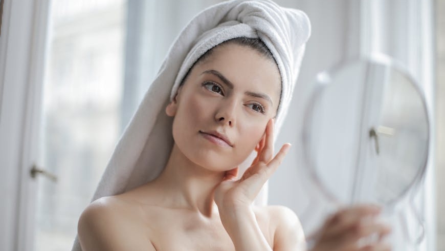 Woman with a white towel around her head, touching her face while looking herself in the mirror.