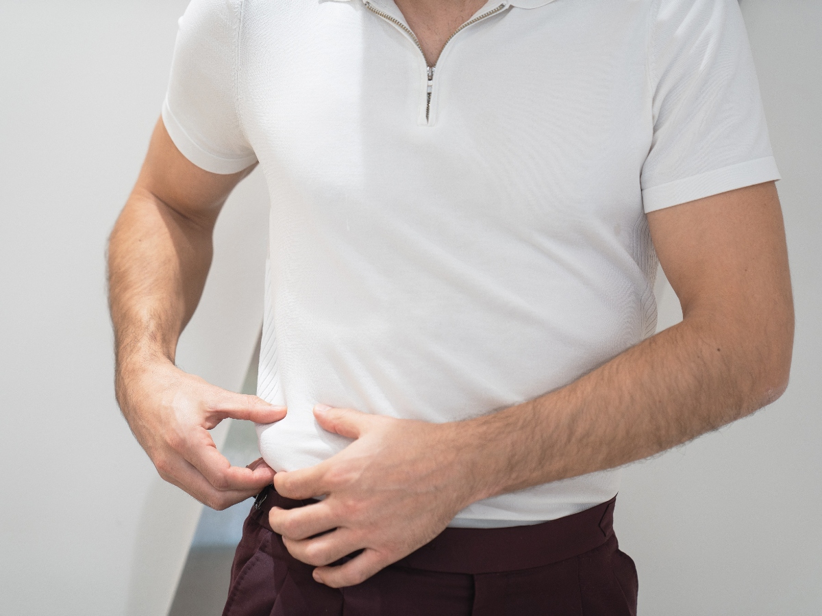 A man adjusts the bottom of his fitted white polo shirt, wearing dark maroon pants, against a light-colored background.